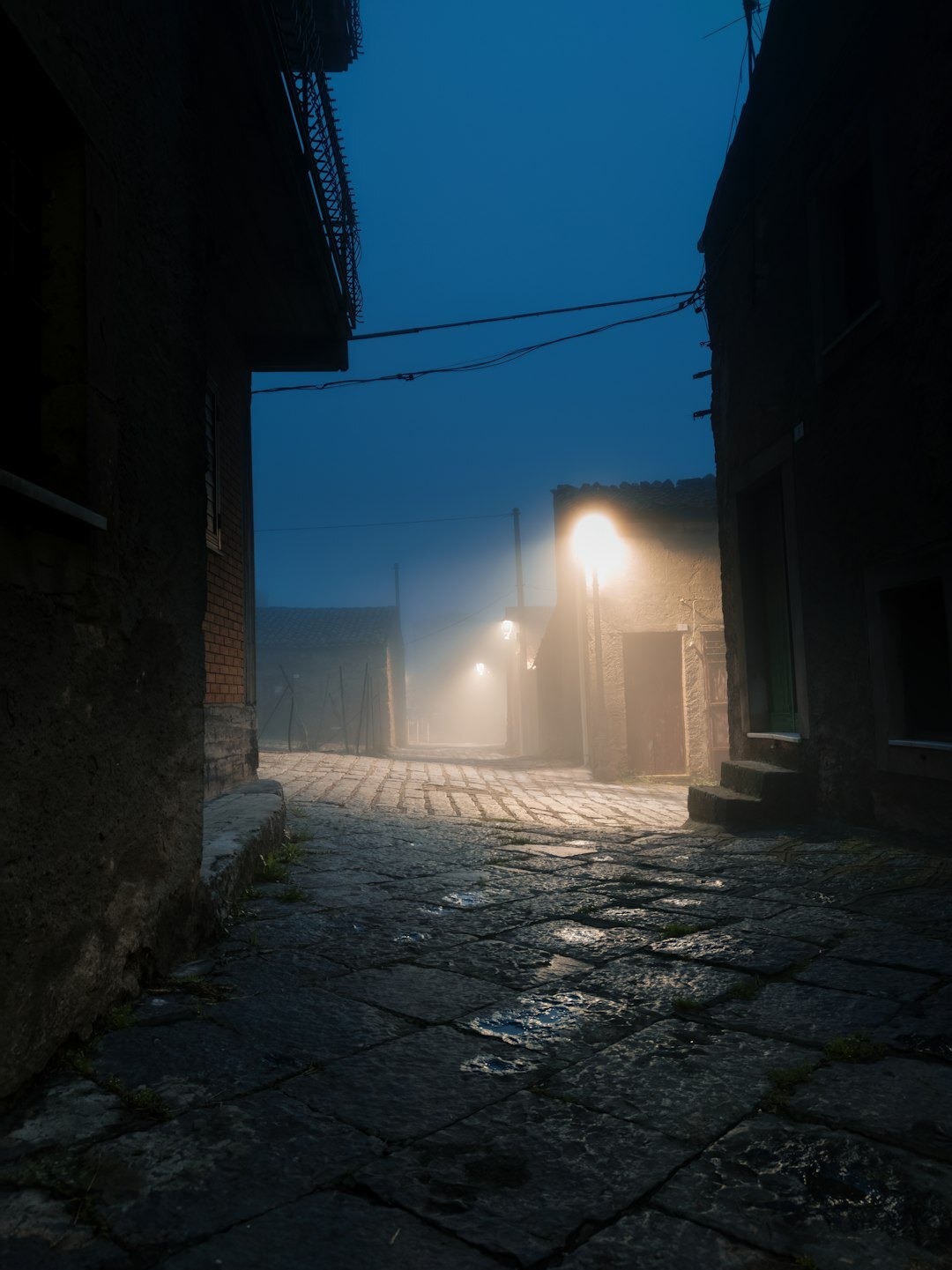 Foggy old European cobblestone street at night with a single lit lamp